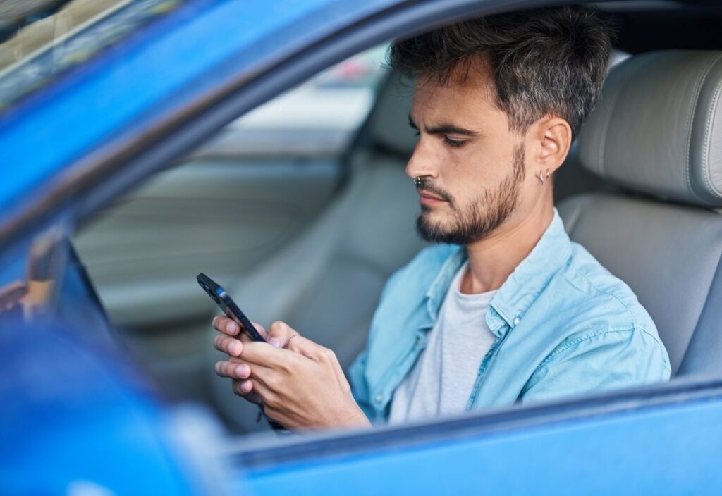 A man sits in the driver's seat of a parked blue car, carscrolling on his smartphone.