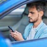 A man sits in the driver's seat of a parked blue car, carscrolling on his smartphone.