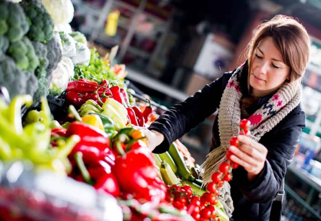 A woman in a coat and scarf selects fresh vegetables, including tomatoes and peppers, at an outdoor market stall.
