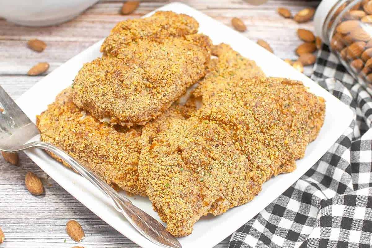 A white plate with several pieces of breaded, baked chicken next to a fork on a wooden table, with a black and white checkered napkin and almonds nearby.