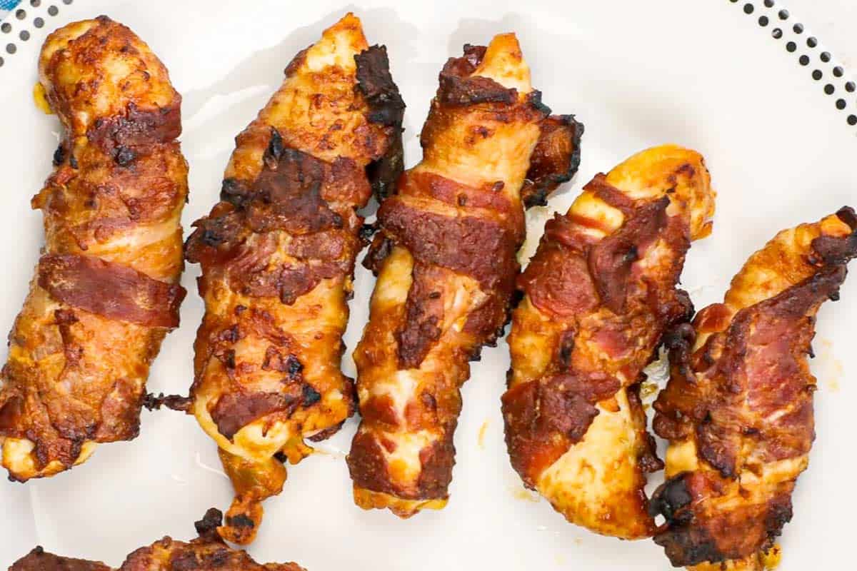 Five pieces of cooked bacon-wrapped chicken are arranged on a white plate with a dotted border.