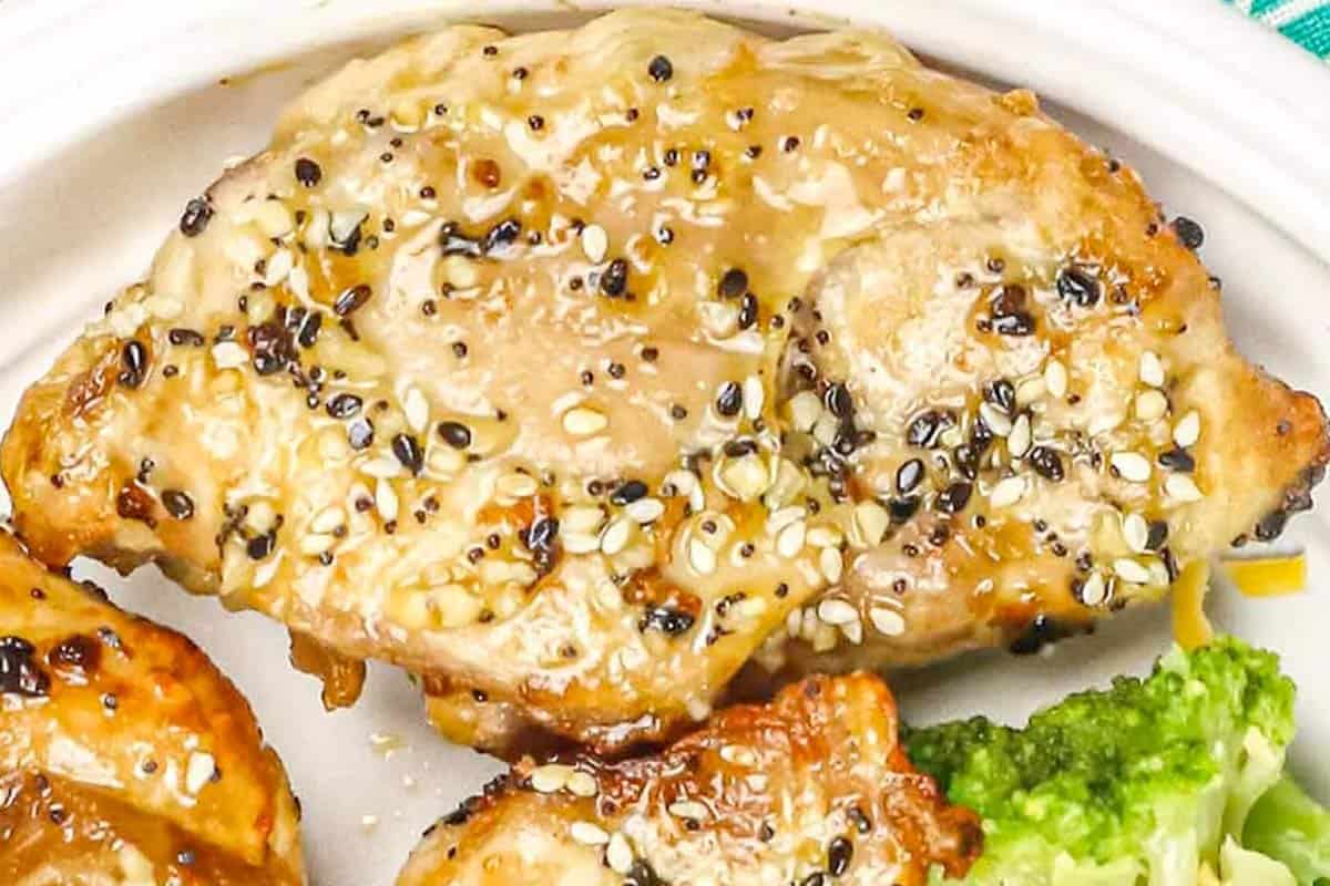 Close-up of a cooked chicken breast topped with sesame seeds and seasoning, served on a white plate with broccoli on the side.