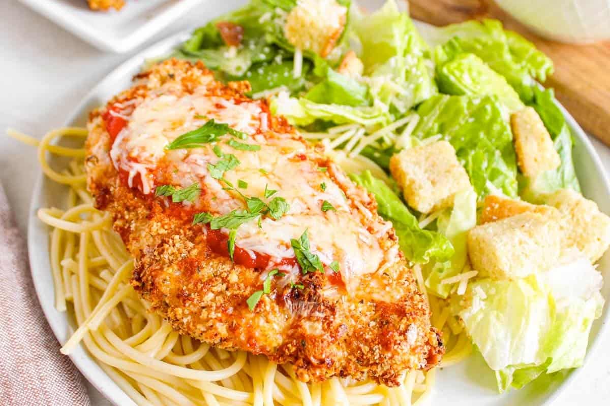A plate of air-fried chicken Parmesan rests on a bed of spaghetti, topped with marinara sauce and cheese, garnished with parsley. Next to it is a Caesar salad with lettuce, croutons, and dressing.