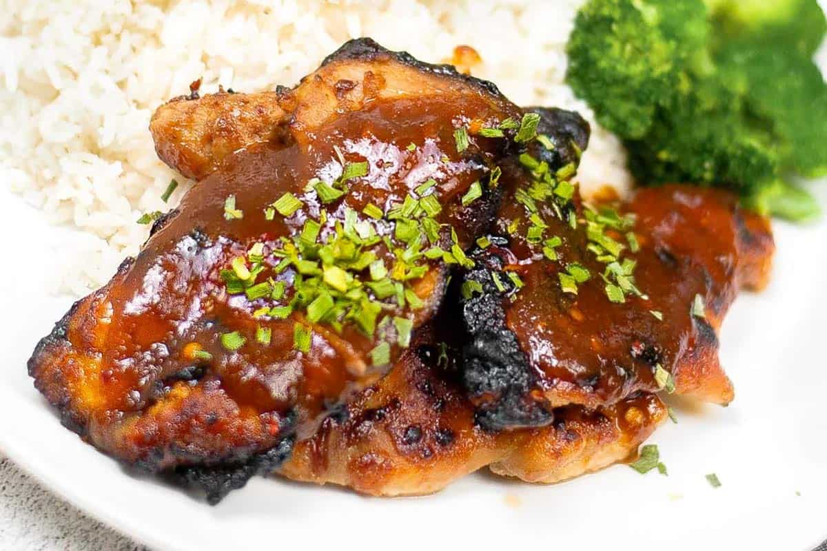 Grilled chicken thighs topped with sauce and chopped herbs, served with white rice and broccoli on a white plate.