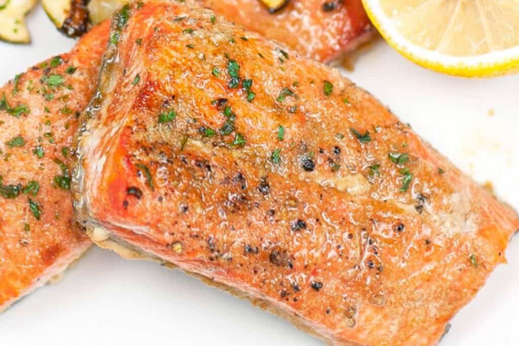 Grilled salmon fillet garnished with herbs and black pepper, served with a lemon slice on a white plate.