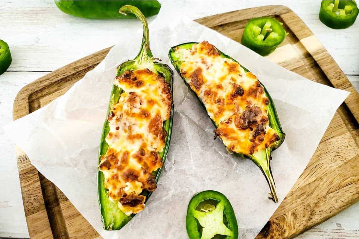 Two baked stuffed jalapeño halves with melted cheese and browned tops, placed on parchment paper atop a wooden cutting board. Sliced jalapeño pieces are nearby.