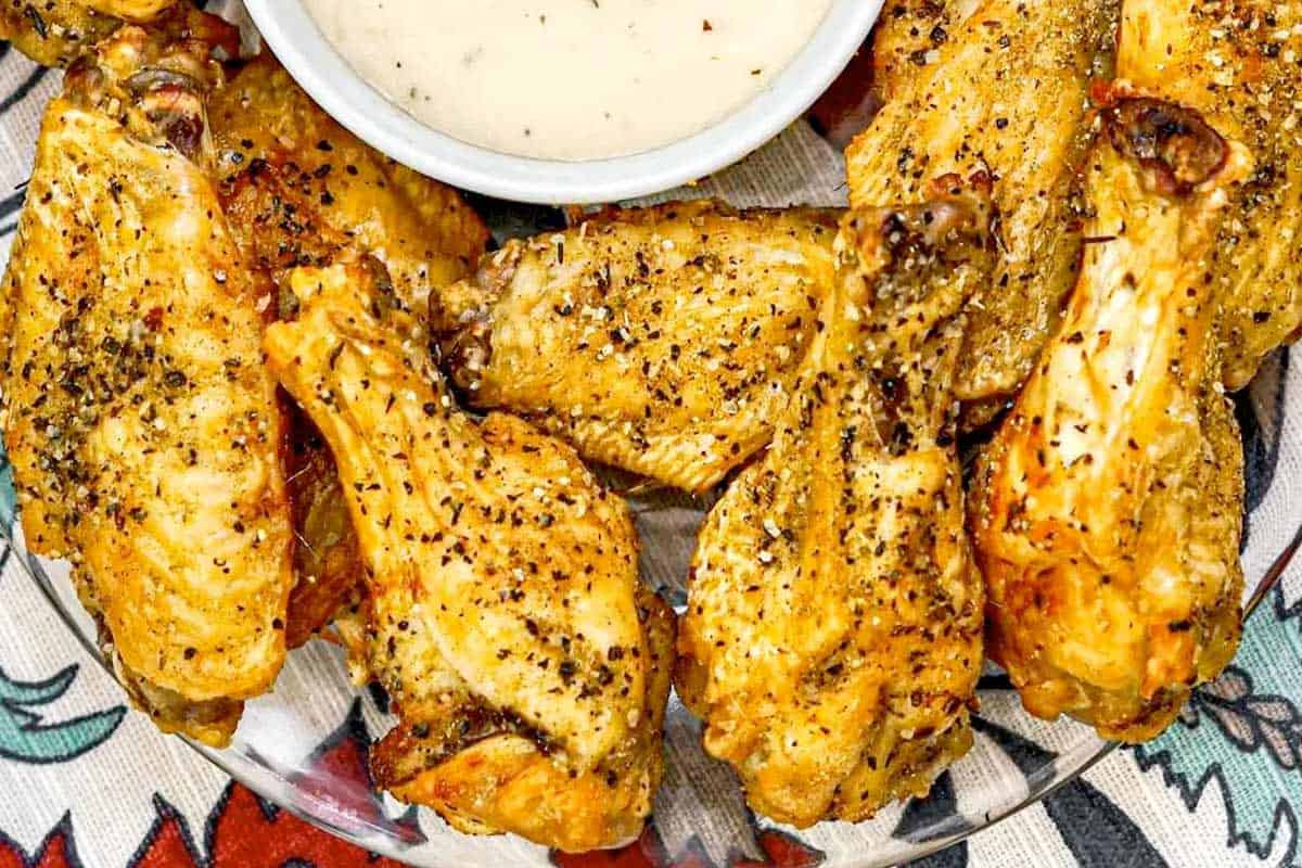 A plate of seasoned chicken wings is arranged next to a bowl of creamy dipping sauce.