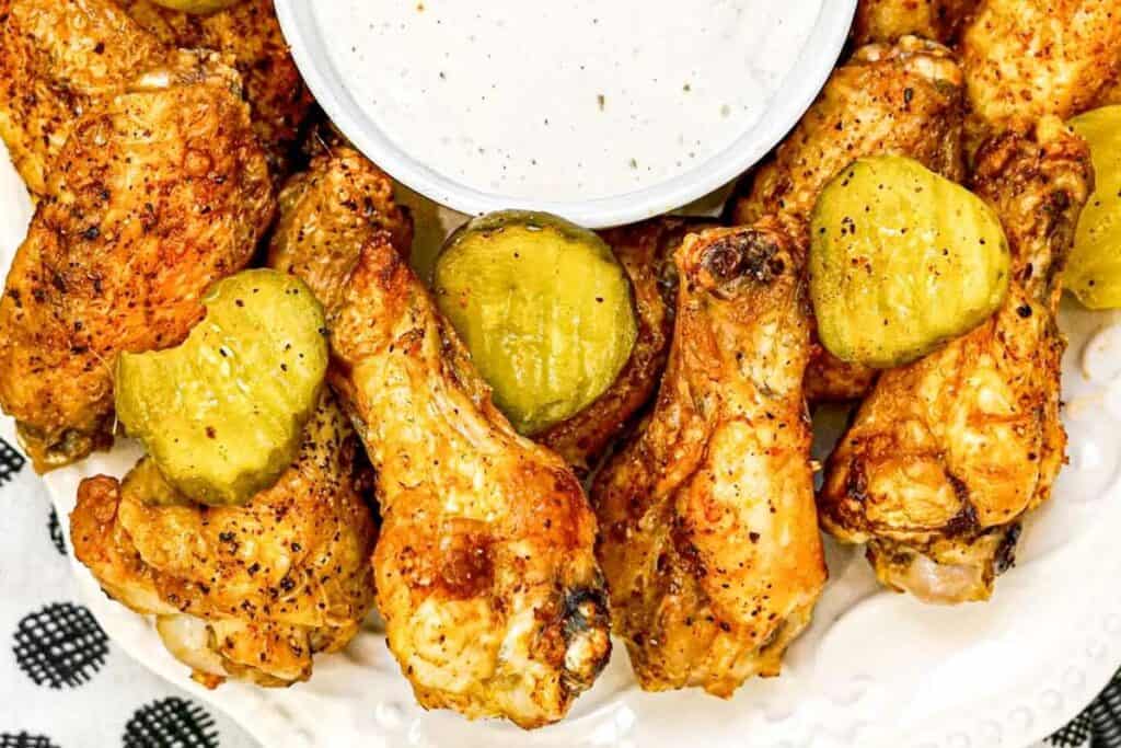 A plate of seasoned chicken wings topped with pickle slices, arranged around a bowl of creamy dipping sauce.
