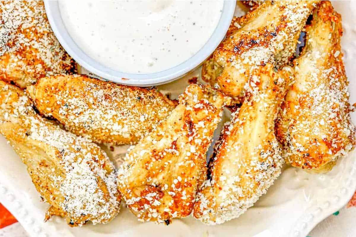 A plate of baked chicken wings coated with grated Parmesan cheese, served with a cup of creamy dipping sauce.