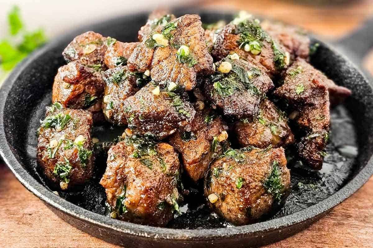 A cast iron skillet filled with cooked, browned steak bites garnished with chopped herbs and minced garlic.
