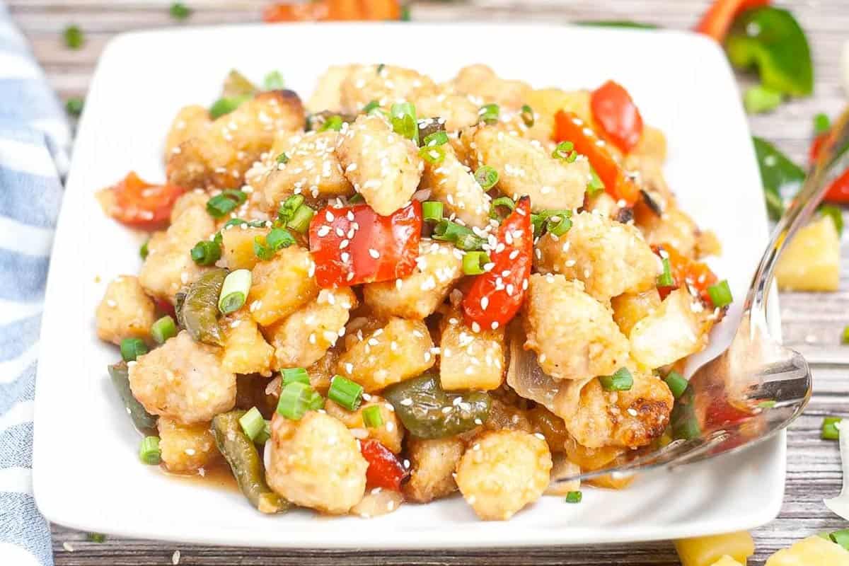 A plate of sweet and sour chicken with bell peppers and pineapple, garnished with chopped green onions and sesame seeds, served with a spoon on a white square dish.