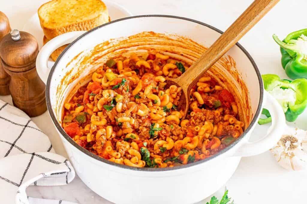 A white pot filled with cooked goulash pasta with ground meat, tomatoes, and herbs, perfect for a comforting weekend dinner. A wooden spoon rests inside, with slices of toast, green peppers, and seasonings nearby.