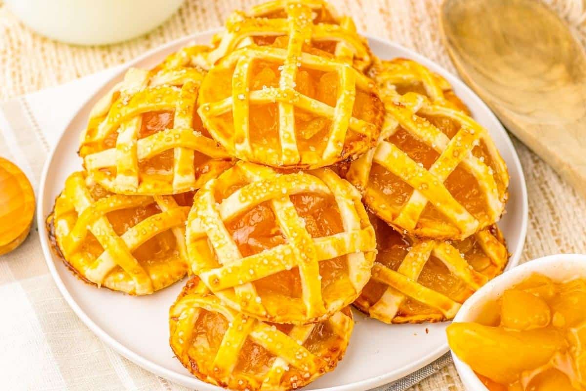 A plate of mini lattice-topped peach pies is displayed on a table next to a wooden spoon and a small bowl of peach filling.