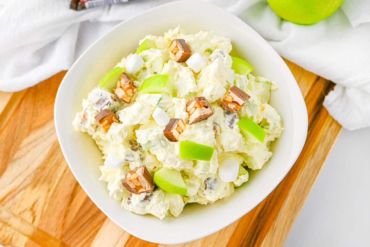 A white bowl filled with a creamy salad topped with apple slices, mini marshmallows, and pieces of chocolate bar, placed on a wooden board.