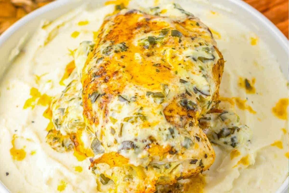 A seasoned, herb-crusted chicken breast topped with creamy sauce, served on a bed of mashed potatoes in a white bowl.