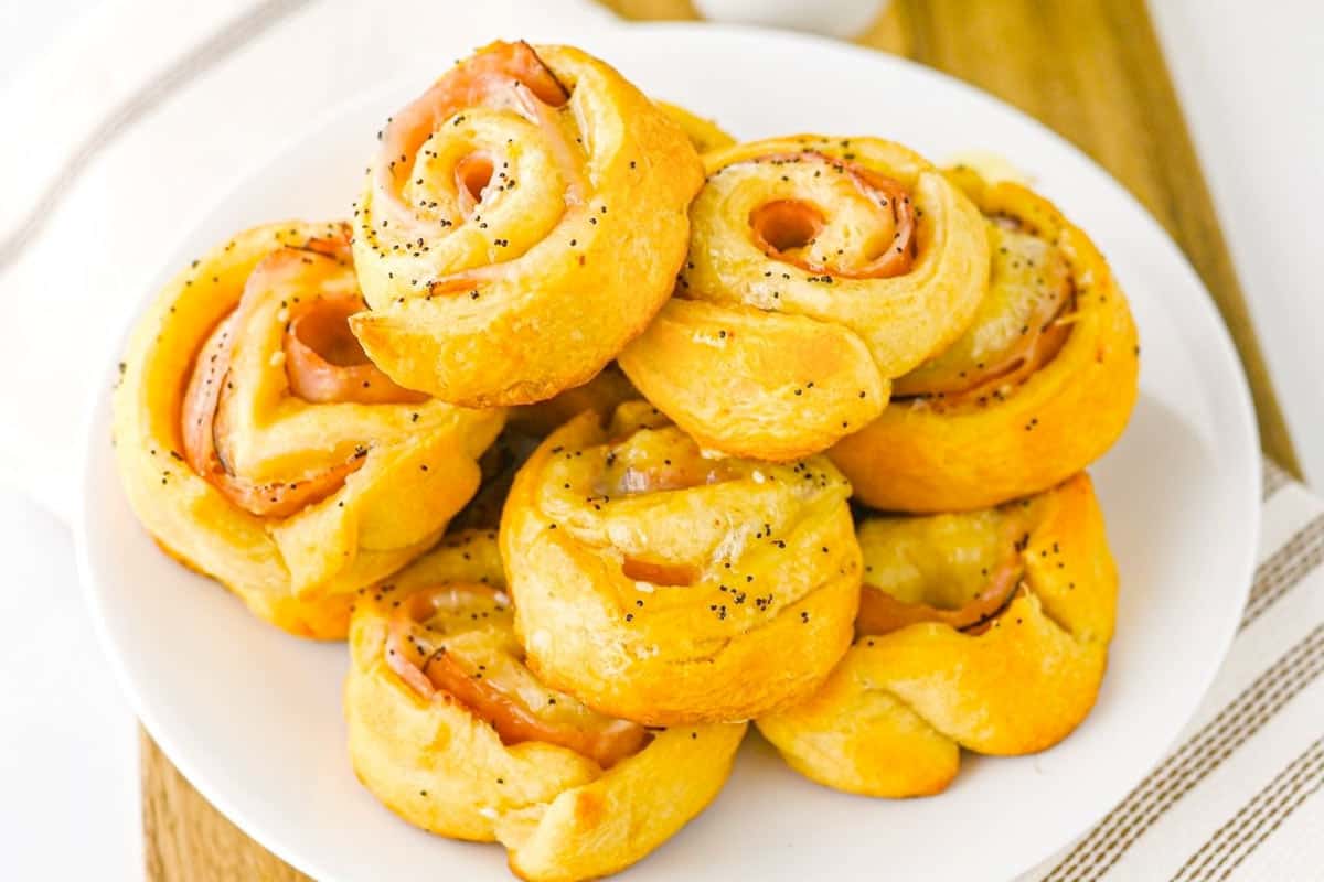 A white plate holds several ham and cheese crescent roll pinwheels topped with poppy seeds, set on a wooden board.