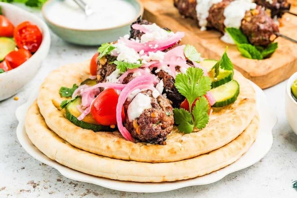 A plate with pita bread topped with juicy grilled meatballs, sliced cucumbers, cherry tomatoes, pickled onions, cilantro, feta cheese, and a drizzle of white sauce&mdash;perfect for fans of tasty beef recipes.