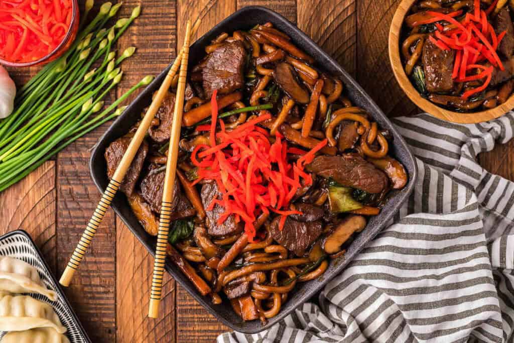 A bowl of stir-fried udon noodles with beef, vegetables, and red pickled ginger, served with chopsticks on a wooden table. Nearby are dumplings and fresh chives&mdash;a delicious example of Asian-inspired dishes.