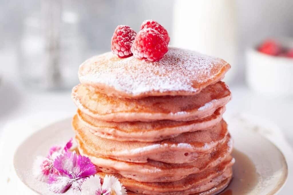 A stack of pancakes topped with powdered sugar and raspberries, garnished with edible flowers on a white plate&mdash;perfect for delightful Valentines Treats.
