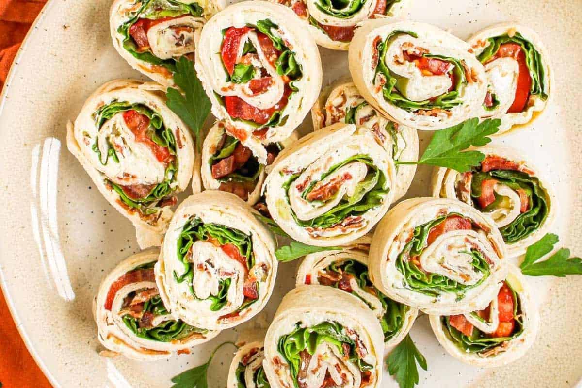 A plate of BLT Pinwheels filled with lettuce, sliced tomatoes, and a creamy spread, garnished with parsley&mdash;an easy snack perfect as a party appetizer.