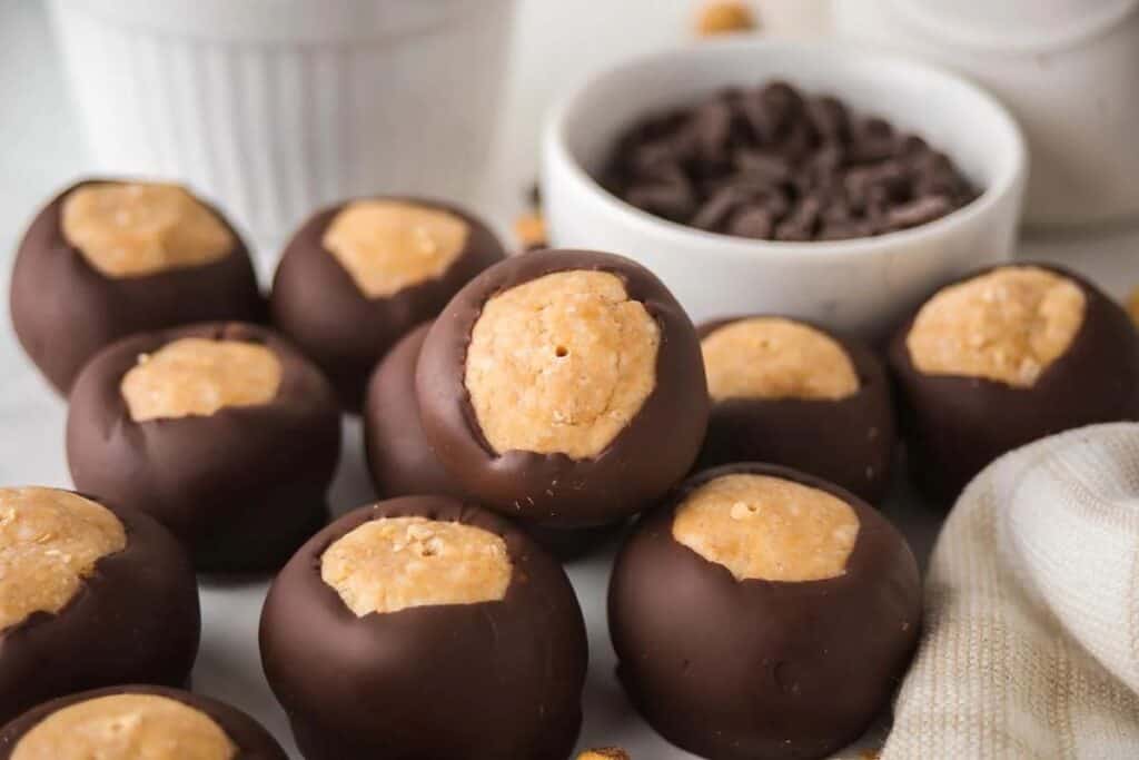 A group of peanut butter balls, classic no bake treats, are partially dipped in chocolate and arranged on a surface with a bowl of chocolate chips in the background.
