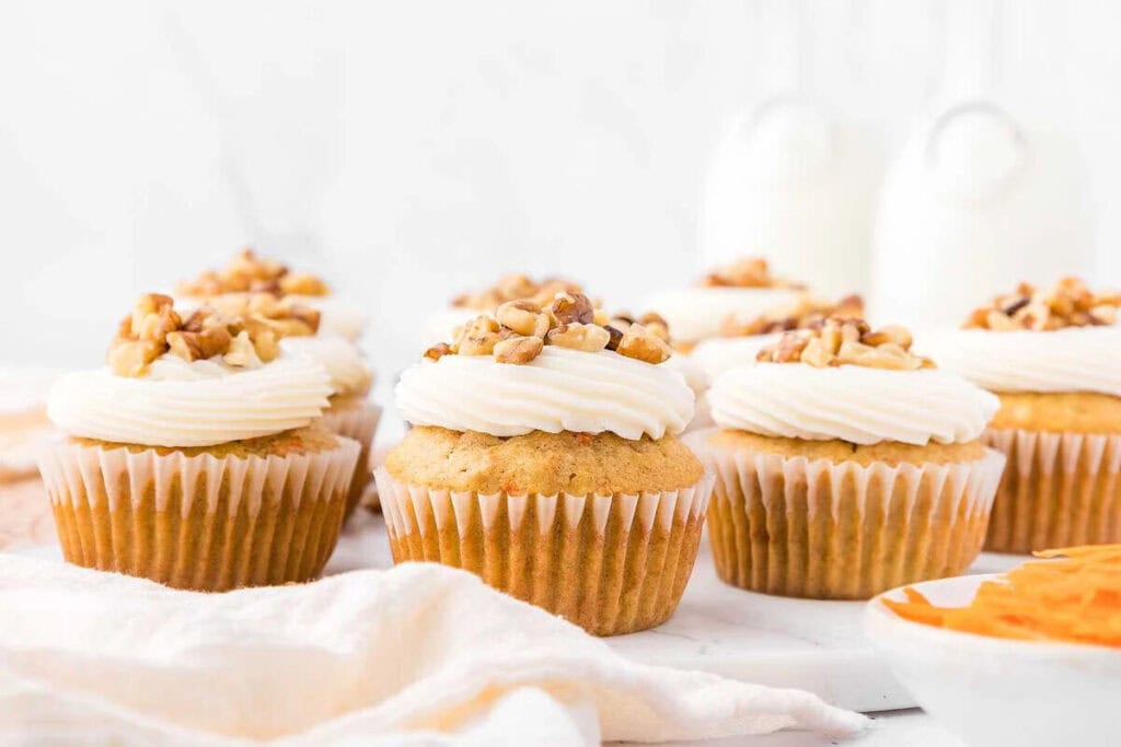 Several cupcakes with white frosting and chopped nuts on top are arranged on a white surface, with milk jars blurred in the background.