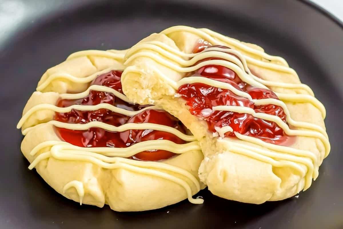 Two shortbread cookies with cherry filling and white icing drizzle are on a black plate, one with a bite taken out of it.
