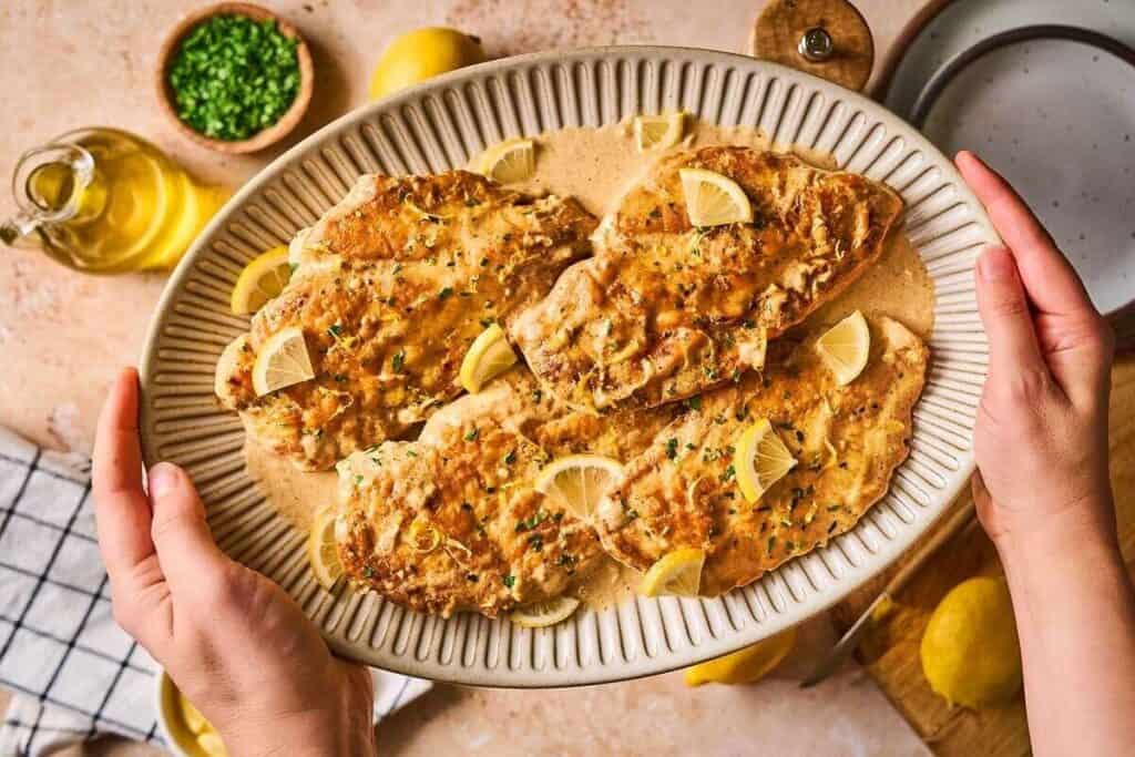 A person holds a platter of cooked chicken breasts in a creamy sauce, garnished with lemon wedges and chopped herbs.