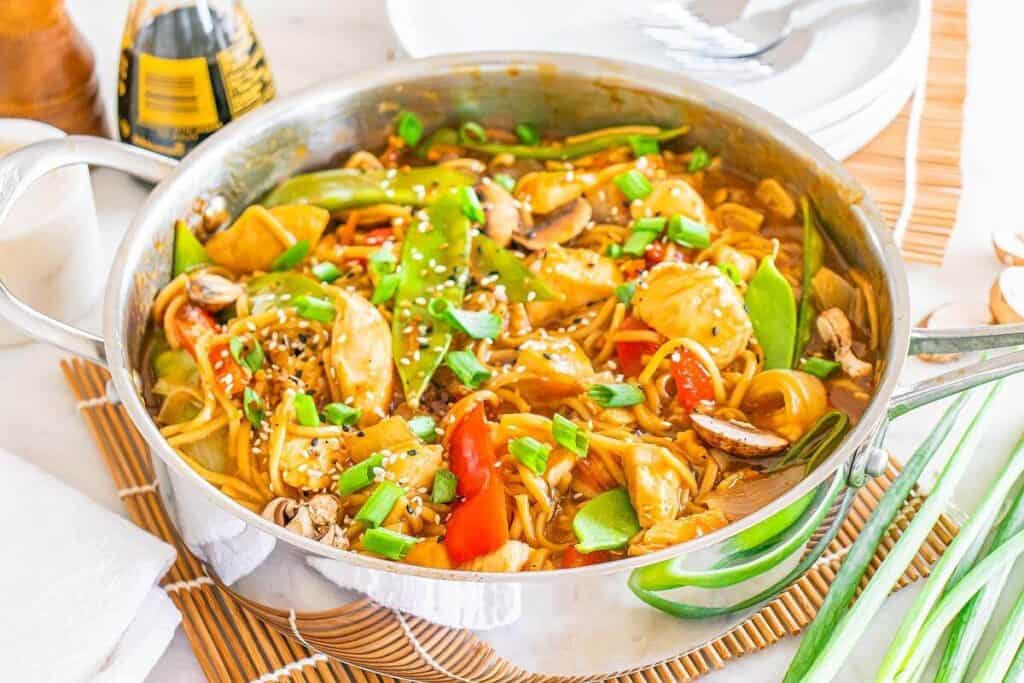 A stainless steel pan filled with stir-fried noodles, vegetables, and tofu showcases Asian-inspired dishes, garnished with green onions and sesame seeds. Plates, chopsticks, and condiments are arranged nearby.