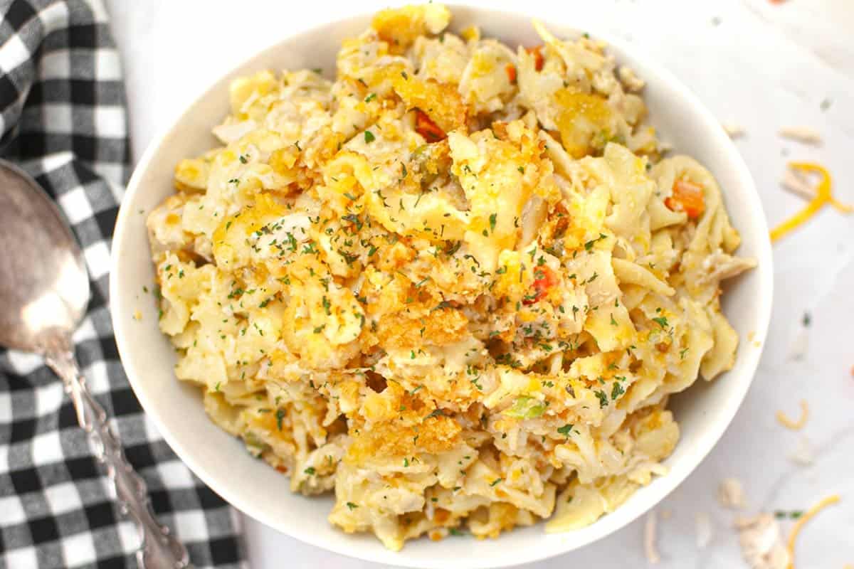 A bowl of creamy pasta casserole topped with breadcrumbs and parsley, placed on a white surface next to a black-and-white checkered napkin and a metal spoon.