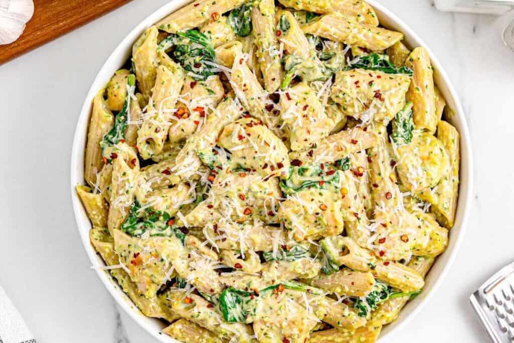 A bowl of penne pasta mixed with creamy sauce, spinach, grated cheese, and red pepper flakes on a white surface.