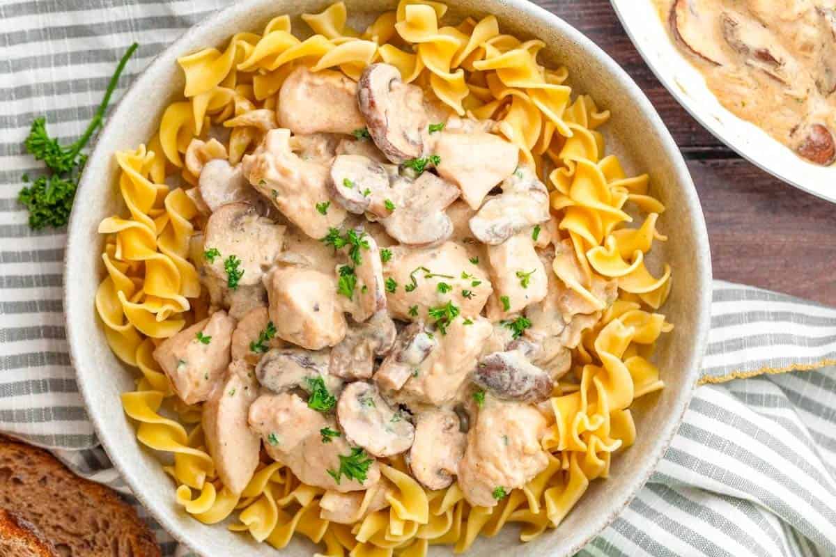 A bowl of egg noodles topped with creamy chicken and mushroom stroganoff, garnished with chopped parsley.
