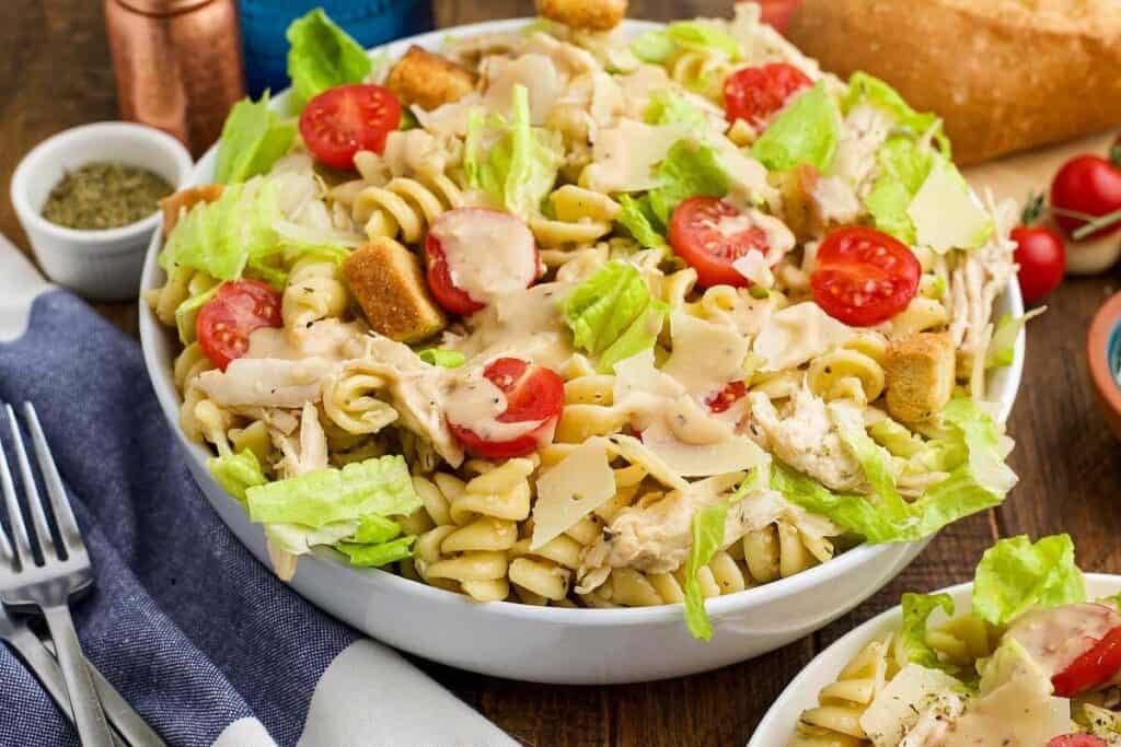 A bowl of Caesar pasta salad with rotini, shredded chicken, lettuce, cherry tomatoes, croutons, and shaved parmesan cheese&mdash;perfect for easy workday meal ideas.