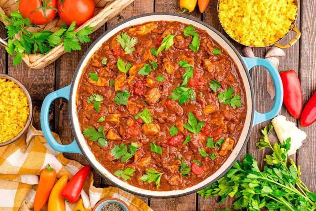 A pot of chicken and tomato stew garnished with parsley, surrounded by bowls of yellow rice, fresh vegetables, and herbs on a wooden table.