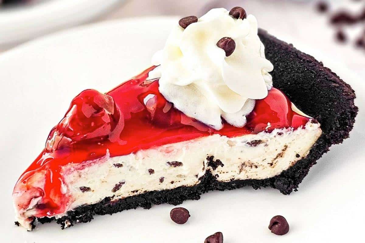 A slice of chocolate chip cheesecake with a dark cookie crust, topped with cherry sauce, whipped cream, and a few chocolate chips on a white plate.