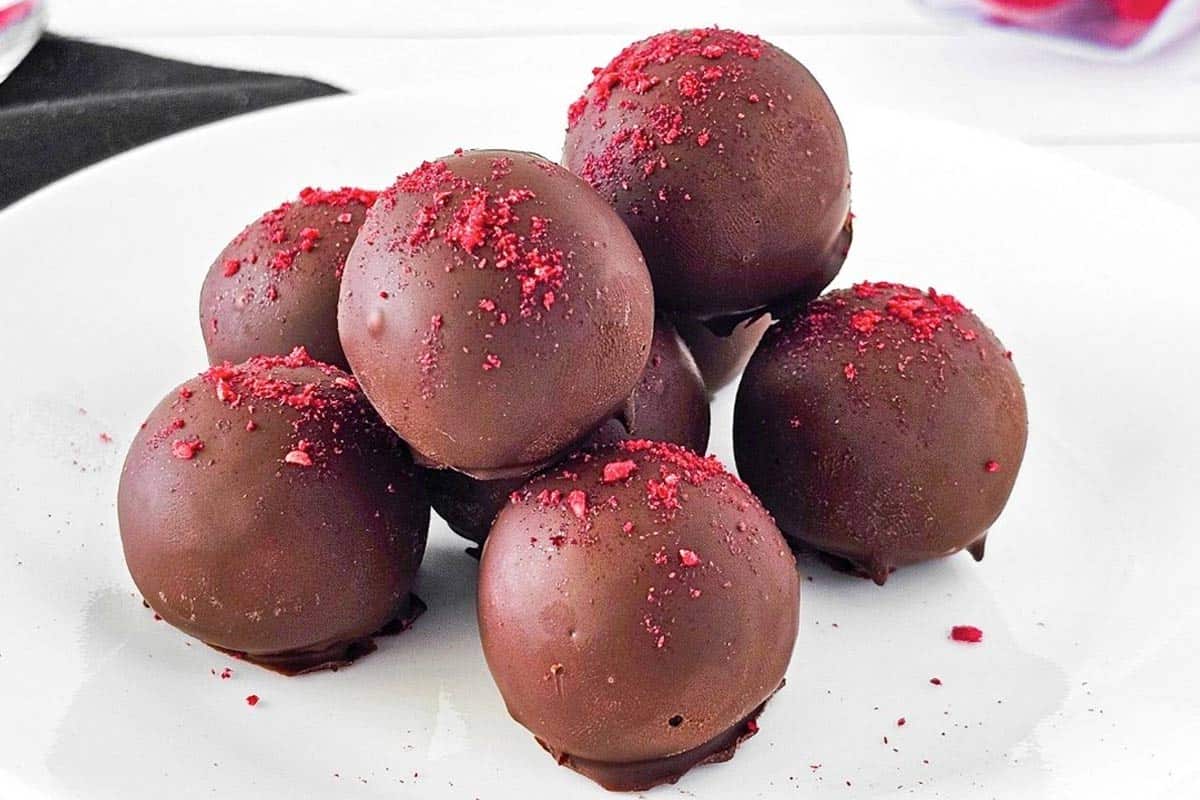 Six round chocolate truffles topped with red powder are stacked on a white plate.