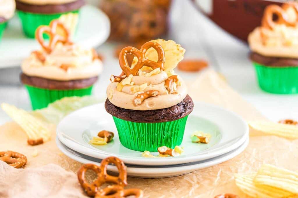 A chocolate cupcake in a green wrapper topped with tan frosting, two pretzels, a potato chip, and pretzel pieces, displayed on stacked white plates.