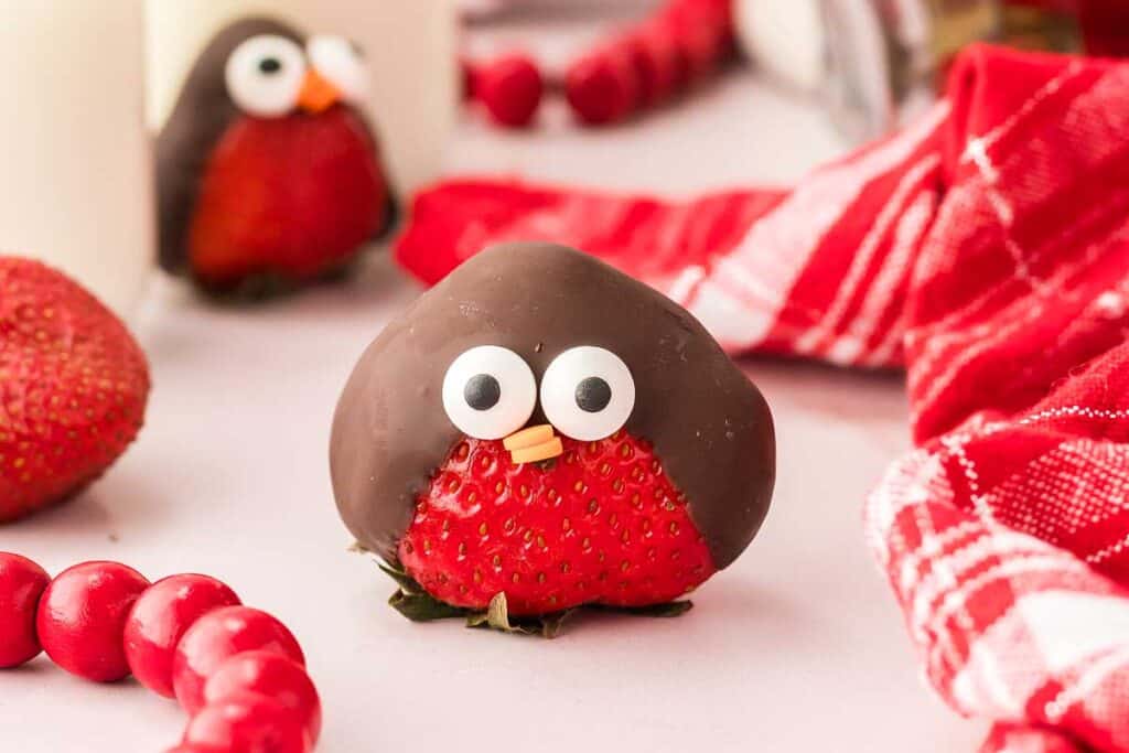 A strawberry partially dipped in chocolate with candy eyes and a small orange candy beak, resembling a bird, is displayed on a table with red decorations—perfect as fun and festive Valentines Treats.