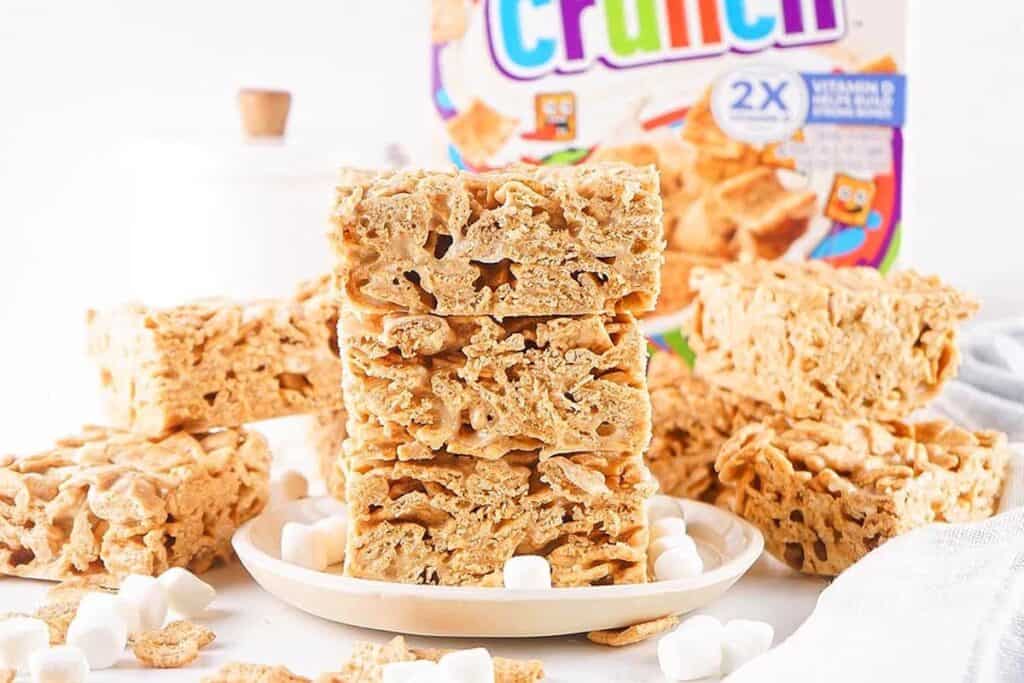 No Bake Treats: Stacked cereal treat bars made with Crunch cereal and marshmallows on a plate, with extra bars, marshmallows, and a cereal box in the background.