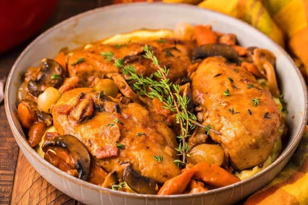 A dish with braised chicken, mushrooms, carrots, onions, and fresh thyme, served in a round bowl on a wooden surface.