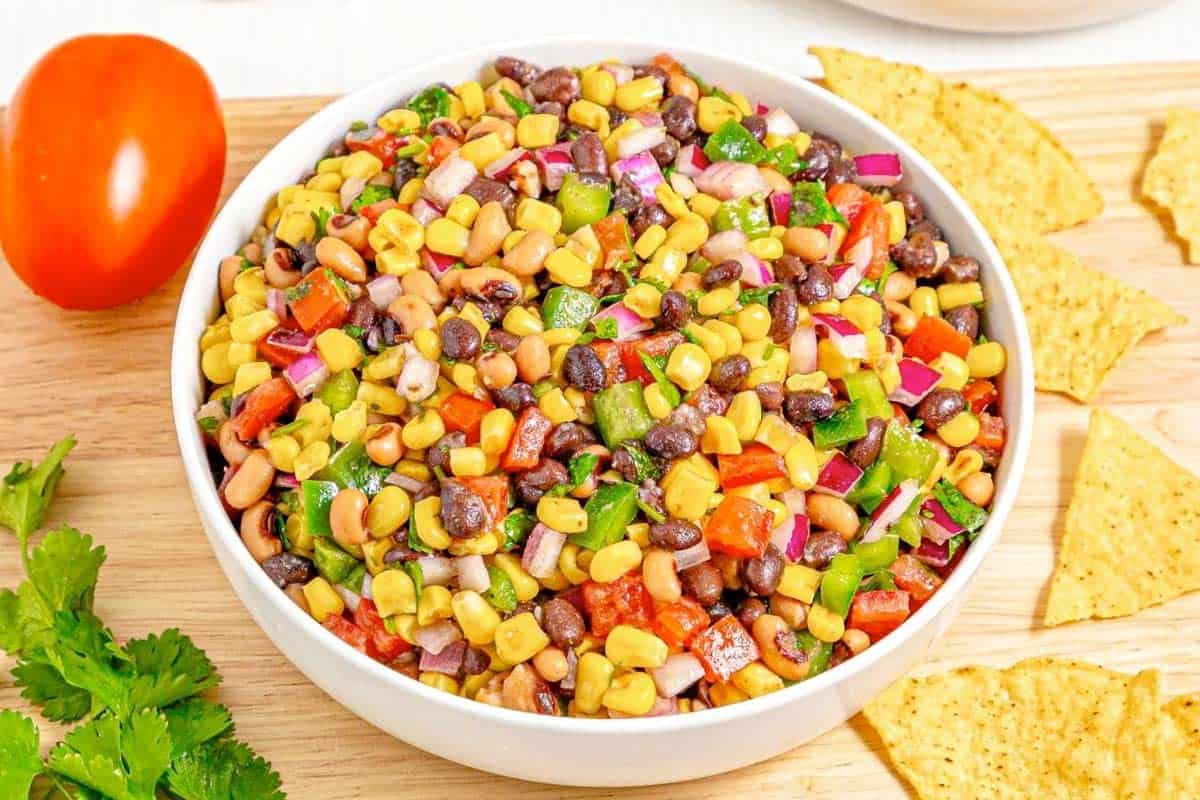 A hand dips a tortilla chip into a bowl filled with Cowboy Caviar, a colorful bean salad of black beans, pinto beans, corn, chopped red onions, green bell peppers, and diced tomatoes.
