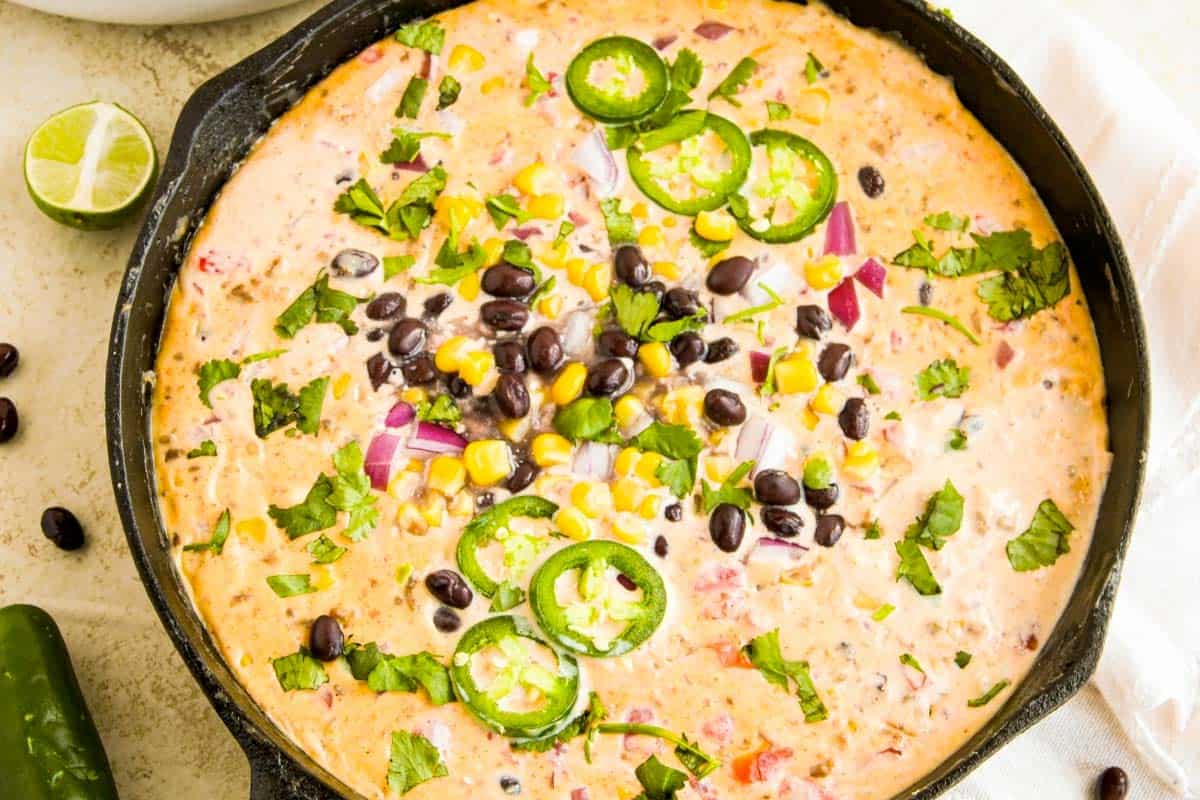 A skillet filled with creamy dip topped with sliced jalapeños, black beans, corn, chopped red onion, and cilantro. A lime wedge and a whole jalapeño are on the surface nearby.