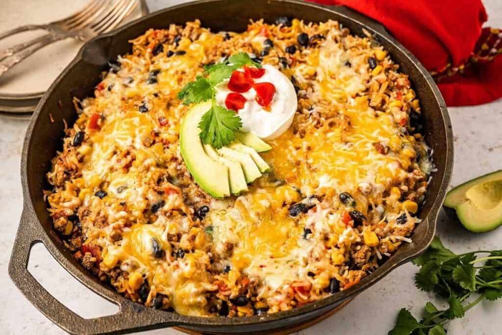 A skillet filled with cheesy rice, black beans, ground meat, and vegetables, topped with avocado slices, sour cream, cilantro, and chopped tomatoes.