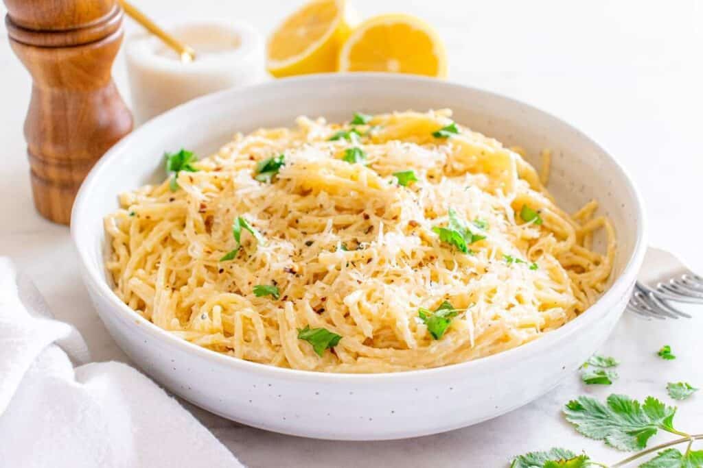 A bowl of creamy spaghetti garnished with grated cheese and fresh herbs, with a pepper grinder, lemon halves, and a sauce dish in the background&mdash;a perfect choice for lovers of quick pasta recipes.