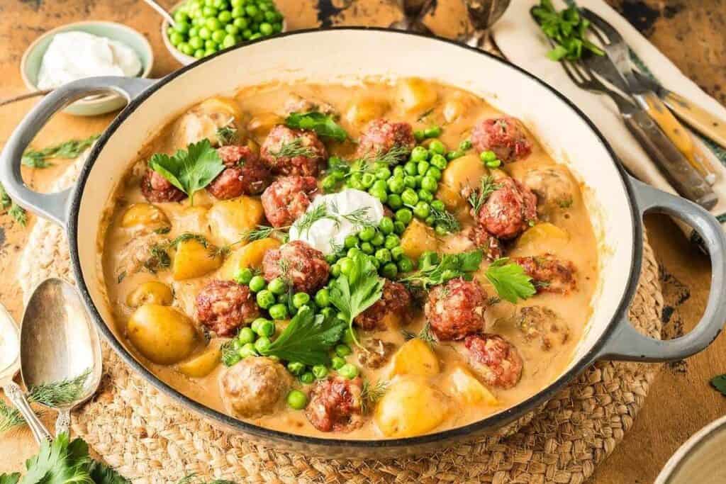 A pan of creamy meatball stew with potatoes and green peas, garnished with parsley and a dollop of sour cream, sits on a woven mat next to utensils and fresh herbs.