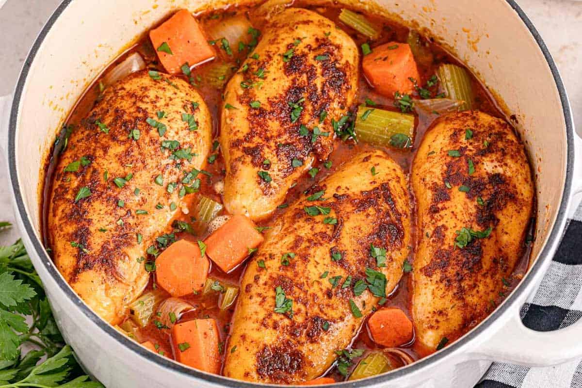 A white pot containing four seasoned chicken breasts, sliced carrots, and celery in a tomato-based broth, garnished with chopped parsley. A bunch of parsley sits nearby.