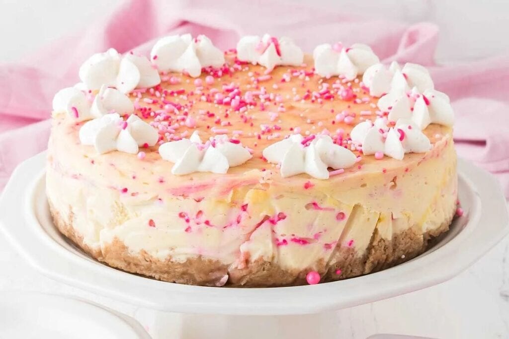 A Simple Cheesecake with a graham cracker crust, topped with whipped cream dollops and pink sprinkles, sits on a white cake stand against a light pink background.
