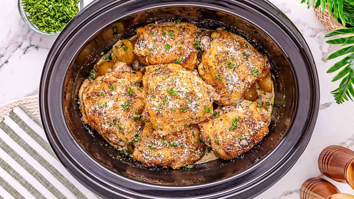 Cooked chicken thighs topped with herbs and grated cheese in a black slow cooker, surrounded by a striped towel, green plant, and small bowl of chopped parsley.