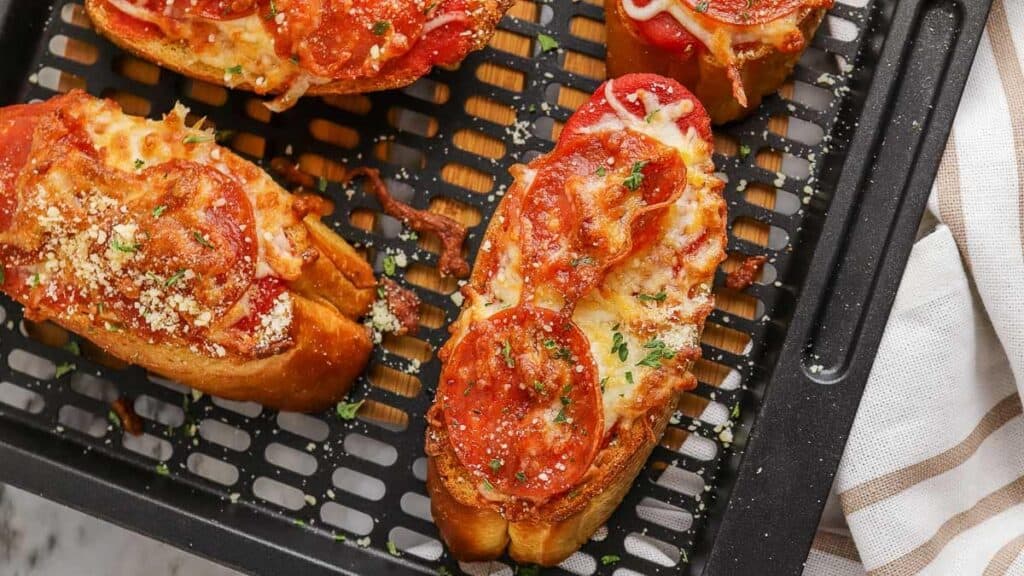 Several slices of toasted bread topped with melted cheese, pepperoni, and herbs sit on a black air fryer tray.
