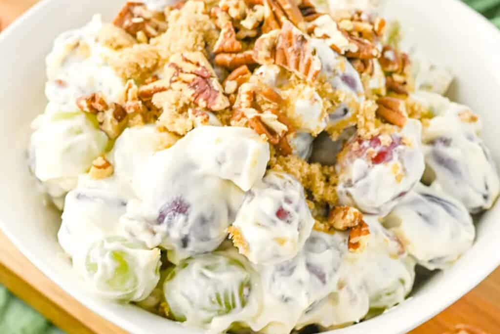 A bowl of grape salad topped with chopped pecans and brown sugar, featuring both green and red grapes coated in a creamy dressing.